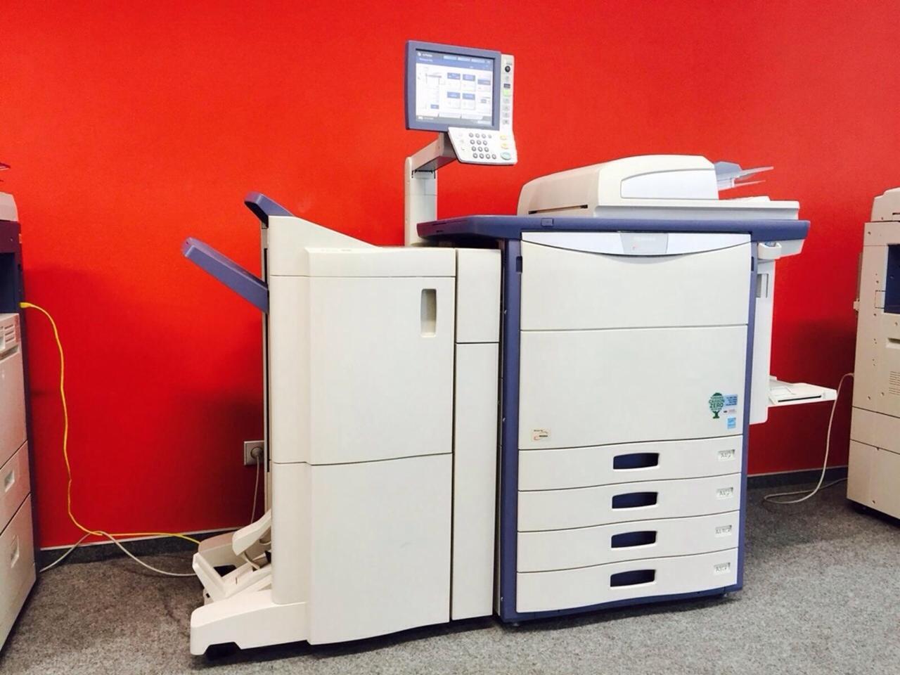 Używana kserokopiarka Toshiba Estudio 5520/5540 cse z panelem dotykowym na tle czerwonej ściany, widoczne inne urządzenia biurowe.