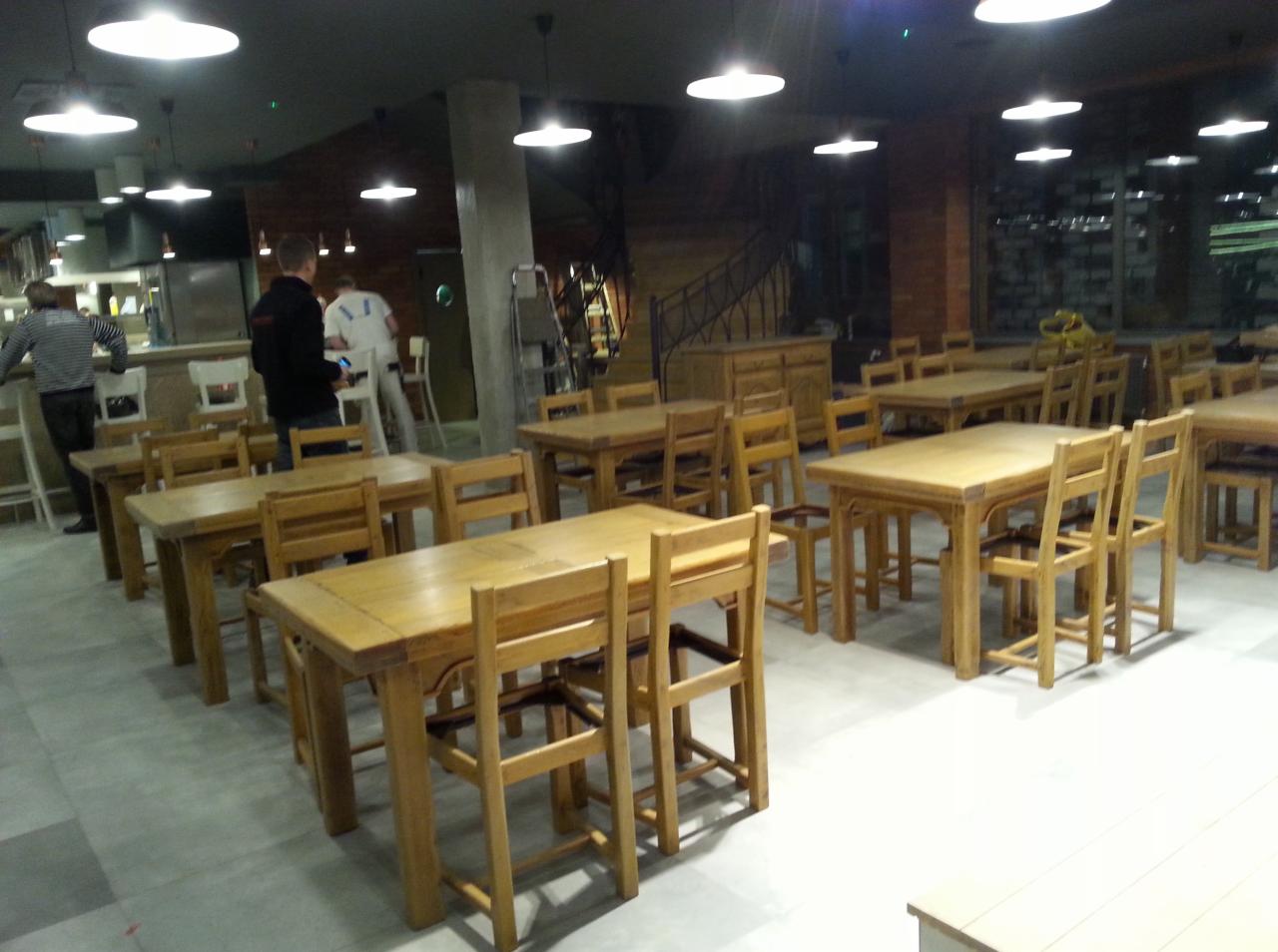 Rustykalne, drewniane stoły i krzesła w jasnym wnętrzu restauracji. W tle widoczny bar i schody. Oświetlenie sufitowe. Kąt ujęcia z perspektywy gościa.