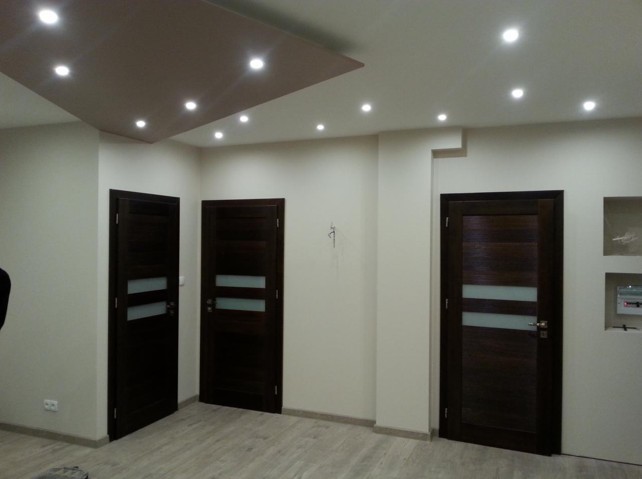 Nowoczesne, ciemne drzwi wewnętrzne z matowymi przeszkleniami, zamontowane w jasnym pomieszczeniu z parkietem i oświetleniem punktowym.