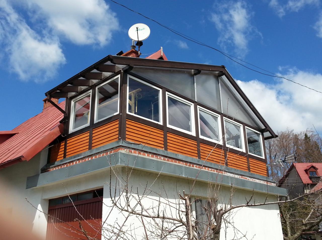 Zadaszony balkon z drewnianą konstrukcją i białymi oknami, widoczny spod kąta. Czerwony dach i antena satelitarna na szczycie budynku. Błękitne niebo z chmurami w tle.
