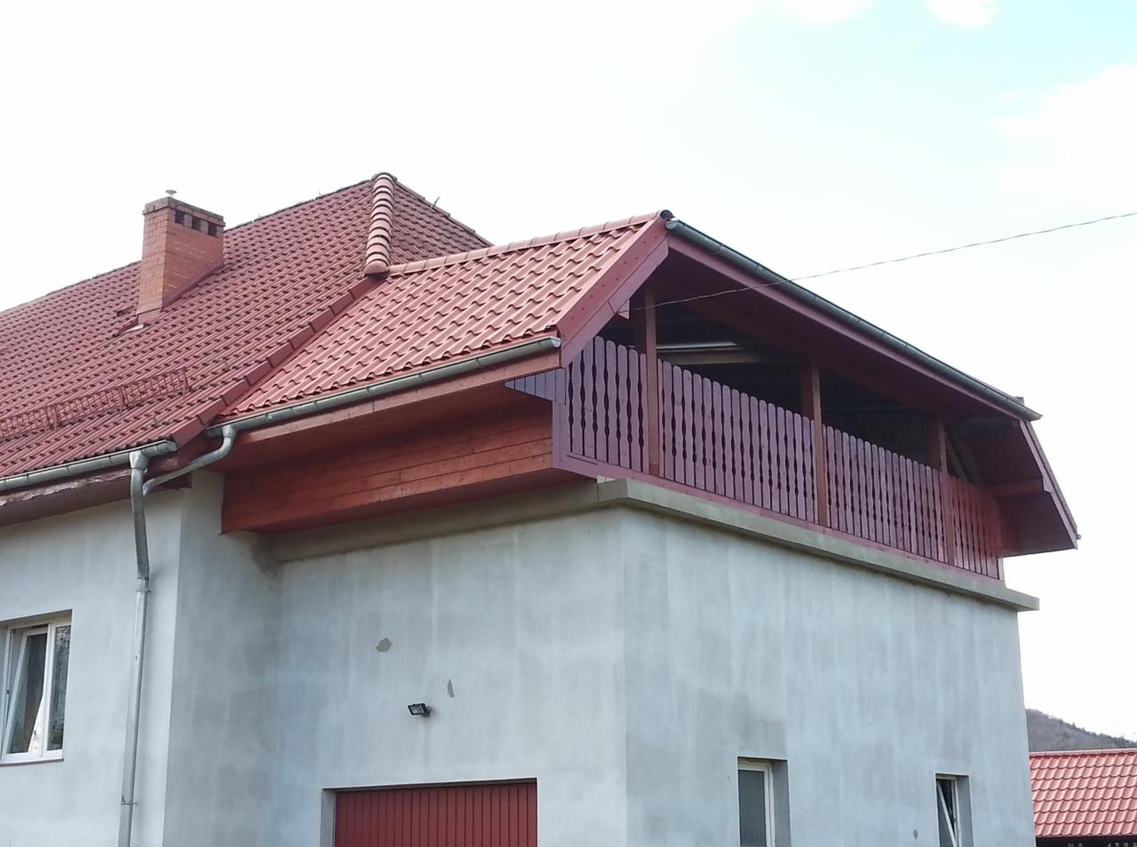 Dom z czerwonym dachem i drewnianą balustradą na balkonie. Widoczne rynny i komin. Ujęcie z dołu, podkreślające konstrukcję budynku i detale wykończenia.