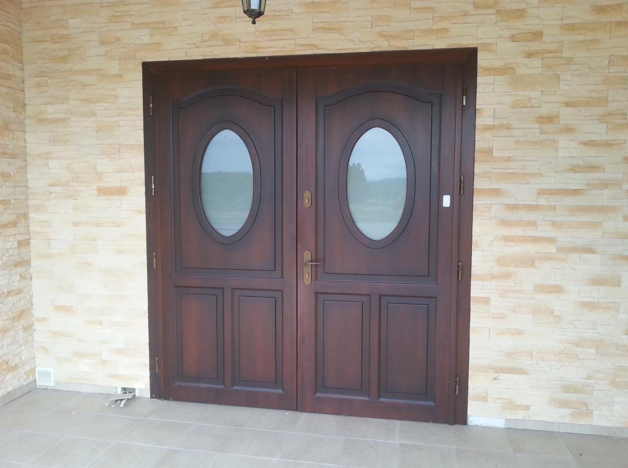 Eleganckie, dwuskrzydłowe drzwi wejściowe z owalnymi przeszkleniami, w ciemnym odcieniu drewna, zamontowane w elewacji z dekoracyjnej cegły.