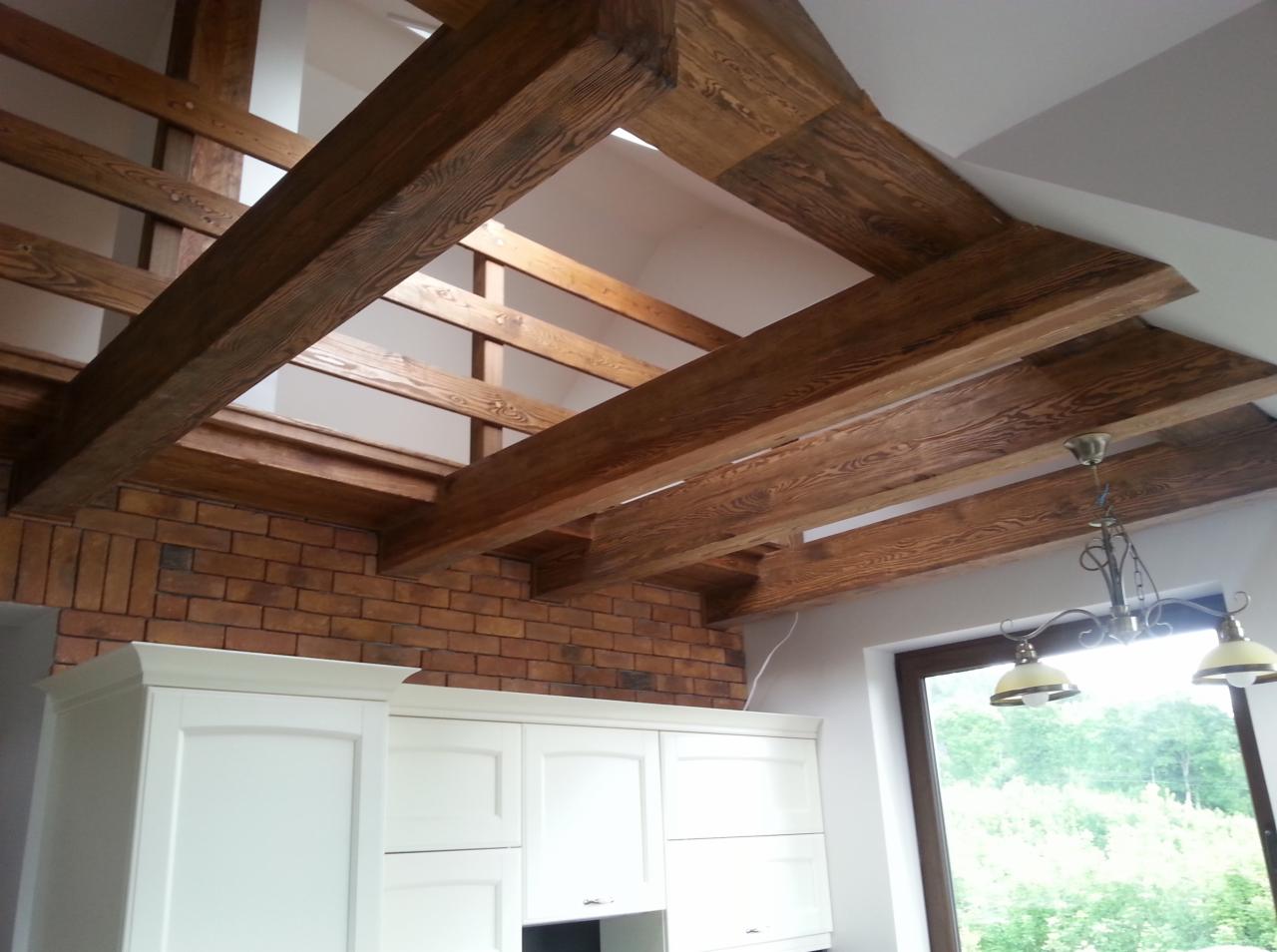 Rustykalne drewniane belki stropowe z widoczną strukturą drewna, ceglana ściana w tle i fragment zabudowy kuchennej w jasnej tonacji.