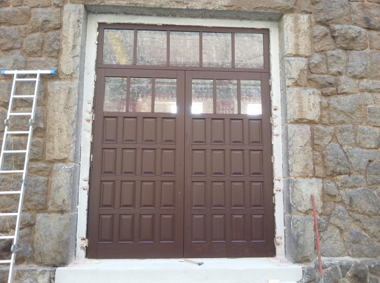 Brązowe, dwuskrzydłowe drzwi z szybami w kamiennej fasadzie budynku. Po lewej drabina, po prawej czerwony pręt. Widoczne ślady prac montażowych.
