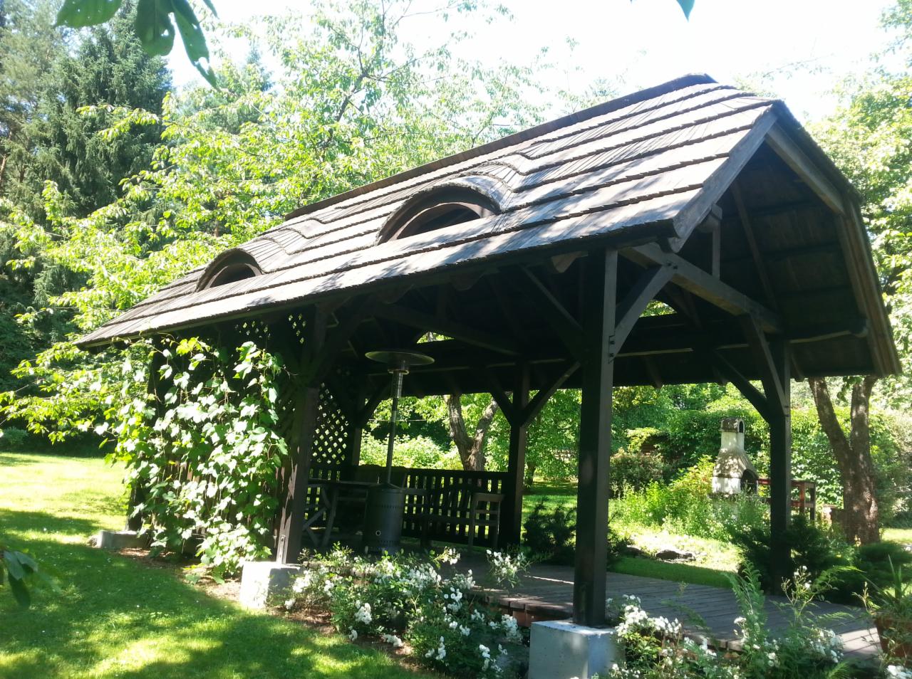 Drewniana altana ogrodowa z unikalnymi, półokrągłymi oknami w dachu, otoczona zielenią i kwitnącymi krzewami, z widocznym paleniskiem w tle.