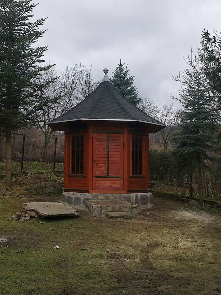 Drewniana sauna ogrodowa z czarnym dachem, umieszczona na kamiennej podstawie, otoczona drzewami iglastymi w pochmurny dzień.