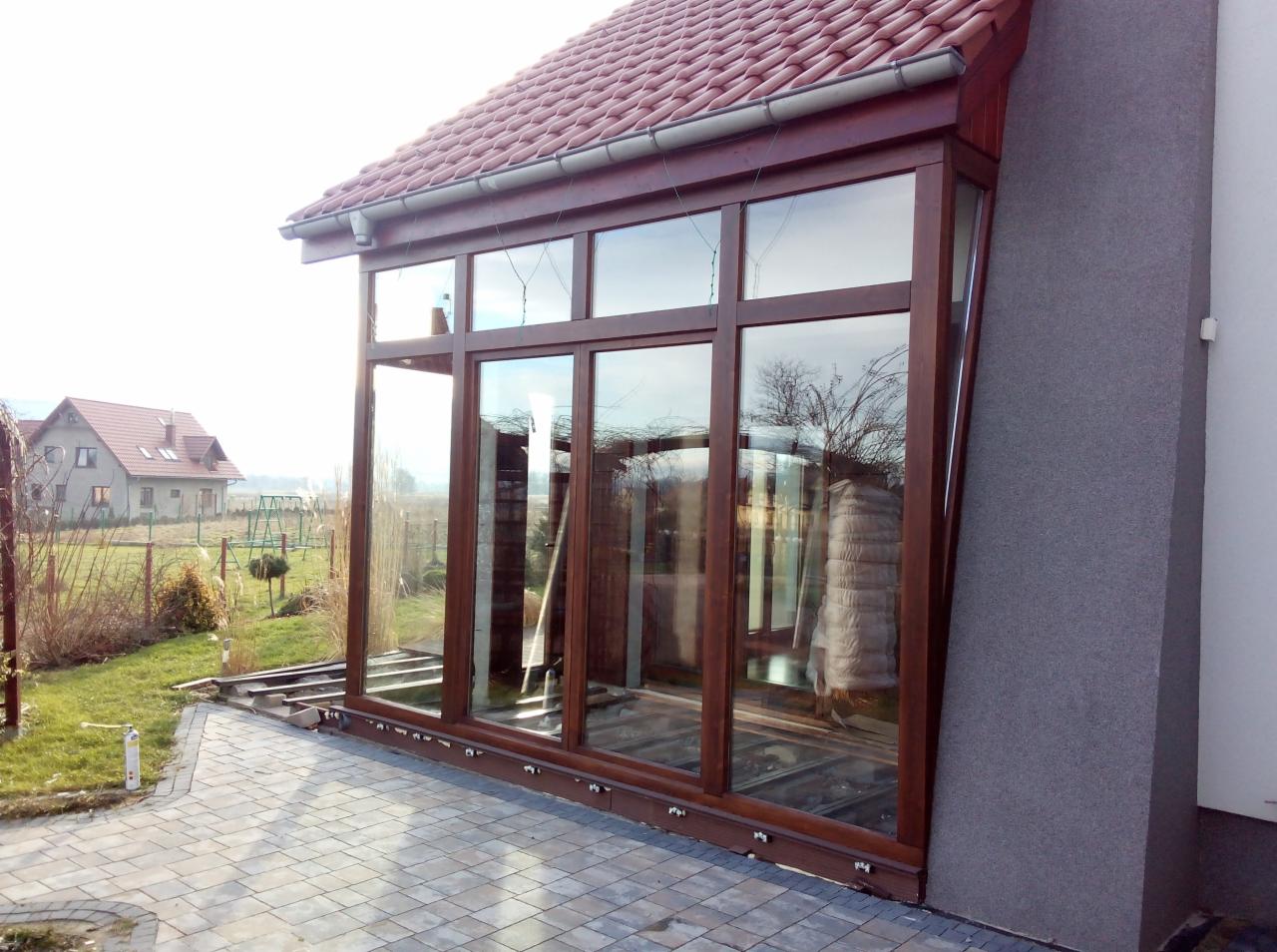 Zewnętrzny widok przeszklonej werandy z drewnianą ramą, przylegającej do domu z czerwoną dachówką. Widoczny fragment ogrodu i kostka brukowa.
