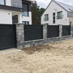 Nowoczesne, poziome ogrodzenie panelowe w kolorze grafitowym z betonowymi słupkami z szarej kostki, otaczające dwie bliźniacze, białe nieruchomości z szarą stolarką okienną.