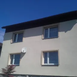 Dwukondygnacyjny dom po remoncie elewacji w beżowym kolorze, z brązowym dachem i białymi oknami. Widoczna antena satelitarna i fragment ogrodu.