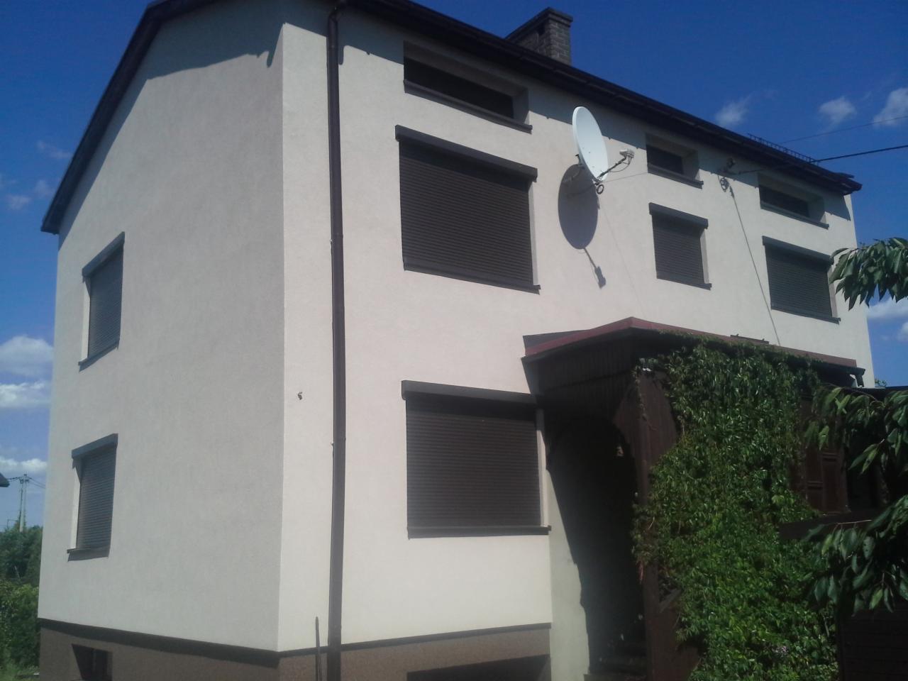 Dwukondygnacyjny dom z elewacją w jasnym odcieniu, brązowe rolety i podmurówka. Biała antena satelitarna na ścianie. Usługi elewacyjne Wodzisław Śląski.