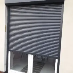 Grafitowa roleta zewnętrzna częściowo opuszczona, odsłaniająca okno PCV z widokiem na wnętrze pomieszczenia z dywanem i krzesłami.