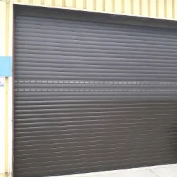 Zewnętrzna, brązowa brama rolowana z poziomymi panelami, zamontowana w budynku przemysłowym z blachy trapezowej.