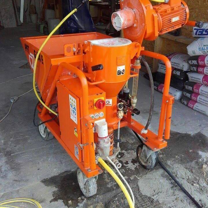 Pomarańczowa maszyna do tynków maszynowych na kółkach, widoczne worki z zaprawą. Sprzęt gotowy do pracy na budowie, detale instalacji i silnika.