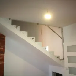 Szklana balustrada schodowa z białymi stopniami i uchwytami ze stali nierdzewnej, widok z dołu, ściana z cegły po lewej stronie.