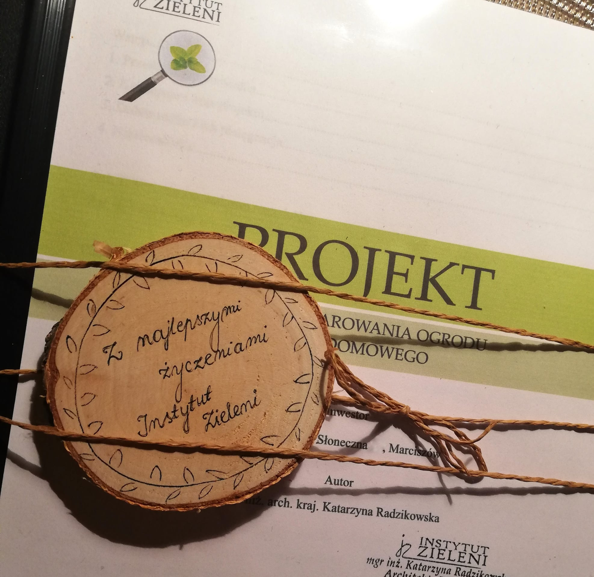 Dokument projektowy z napisem 'Projekt' i adnotacją 'Z najlepszymi życzeniami Instytut Zieleni', ozdobiony drewnianym krążkiem z rysunkami roślinnymi i sznurkiem.