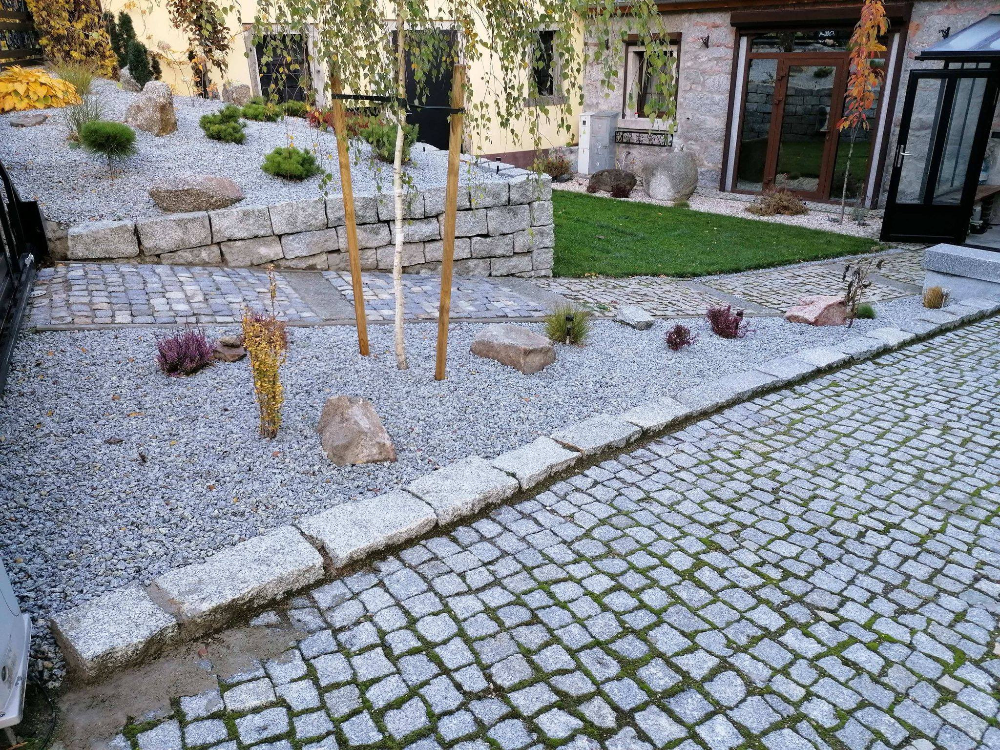 Ogród z kamienną ścieżką, rabatą z kamieni i roślin oraz podtrzymywanymi młodymi brzozami, widok na dom z kamienną elewacją.