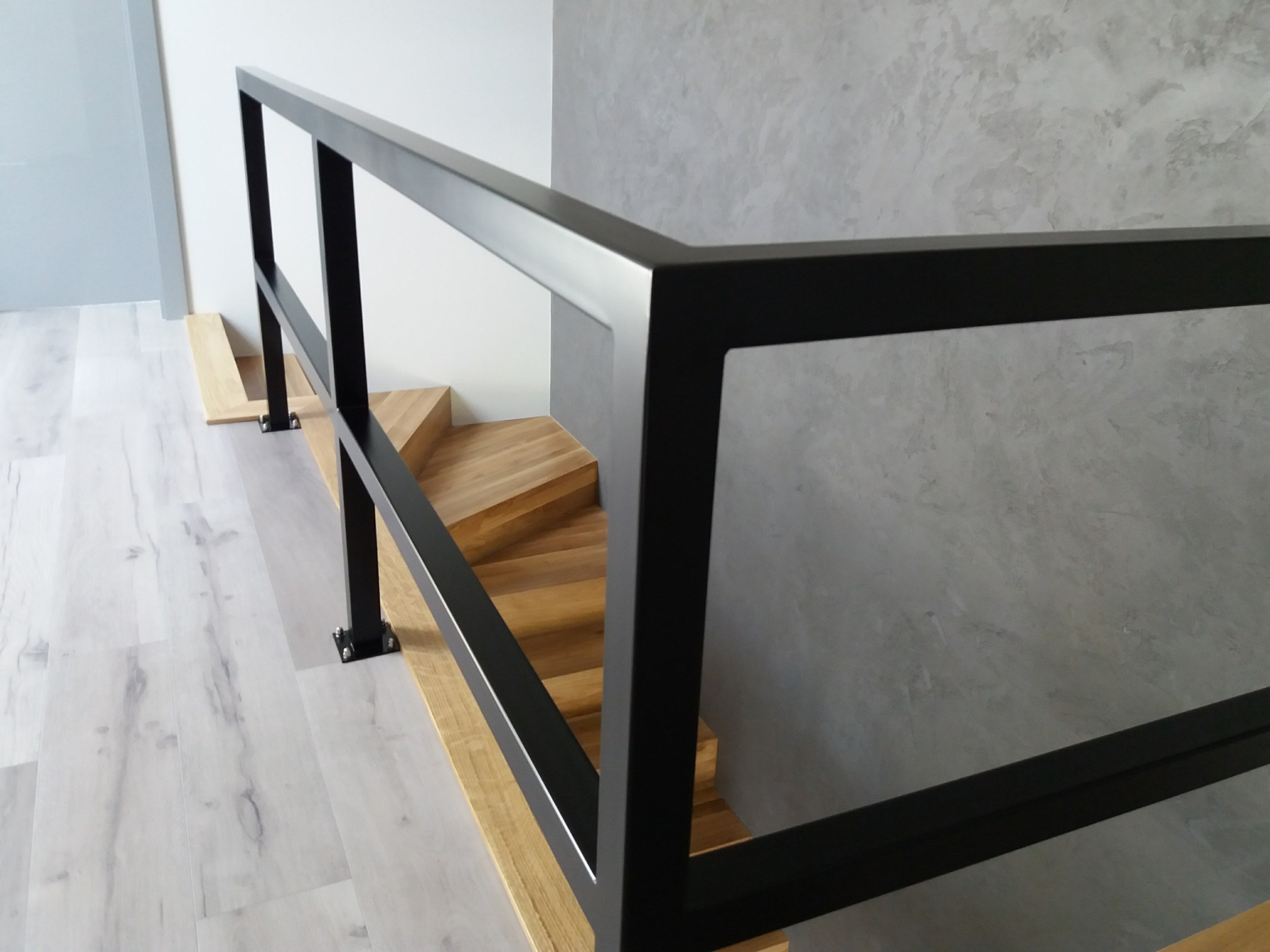 Nowoczesna, czarna barierka schodowa z kwadratowych profili, zamocowana do drewnianych stopni spiralnych schodów, z widoczną fakturą szarej ściany w tle.