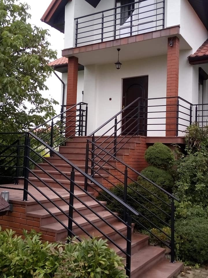 Zewnętrzne schody wejściowe do domu z czarnymi, poziomymi balustradami i ceglanymi elementami wykończeniowymi.
