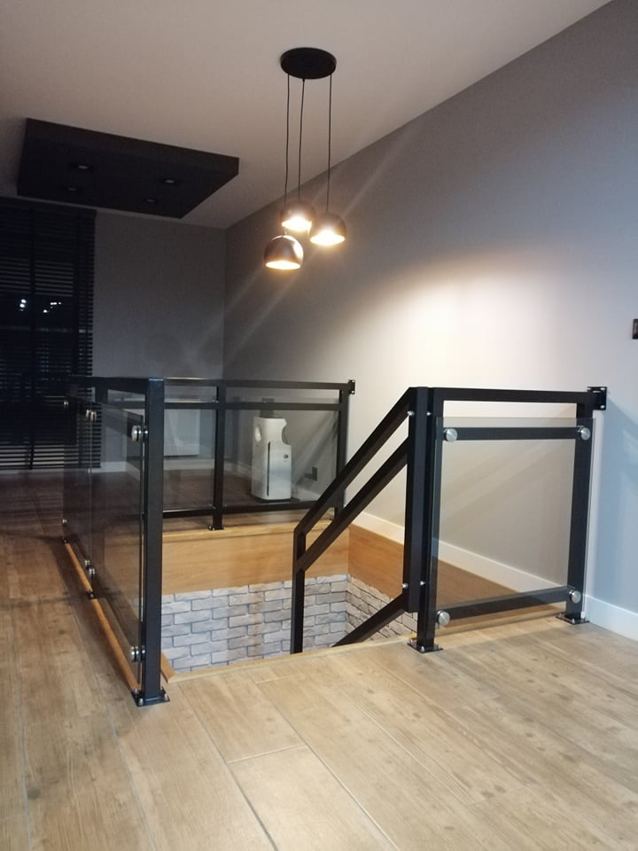 Szklana balustrada schodowa z czarną ramą, widoczna z góry, w domu w Ostrowcu Świętokrzyskim; podświetlona przez wiszące lampy kuliste.