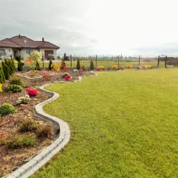 Zadbany ogród z równo przystrzyżonym trawnikiem i rabatami kwiatowymi oddzielonymi kamiennym obrzeżem, na tle domu jednorodzinnego w słoneczny dzień.