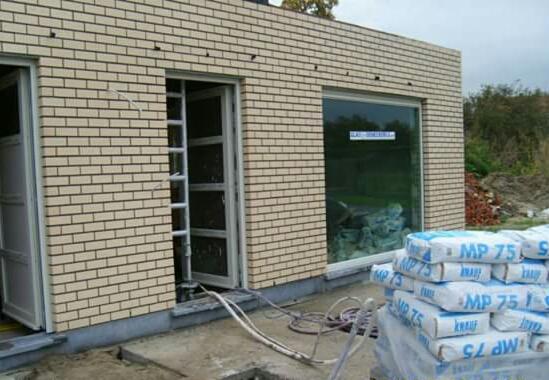 Fragment ściany z cegły klinkierowej w trakcie prac budowlanych. Widoczne okno, drzwi, worki z zaprawą MP 75 i węże do tynkowania maszynowego.