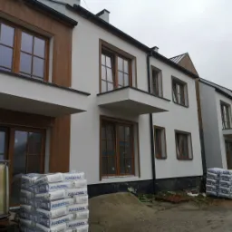 Ocieplona elewacja szeregowca z drewnianymi oknami, widoczne worki z zaprawą Kreisel i zbiornik na wodę, teren budowy w trakcie prac wykończeniowych.