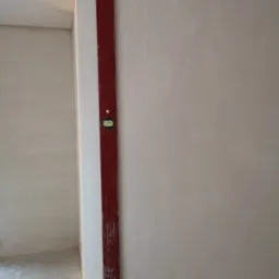 Czerwona poziomica przyłożona do świeżo nałożonej, jasnej ściany, sprawdzana przez osobę w rękawicy; surowy stan wykończenia wnętrza w tle.