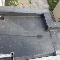 Widok z góry na płaski dach pokryty czarną papą, z widocznymi elementami odwodnienia i fragmentem drabiny w rogu kadru.