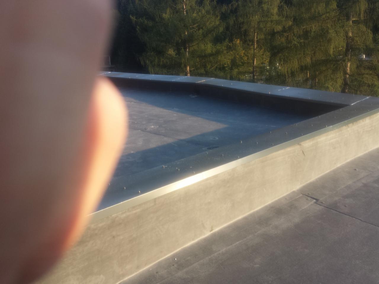 Nowe, płaskie pokrycie dachu z blachy, zabezpieczone metalowym obramowaniem; w rogu kadru widoczny fragment dłoni.