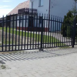 Czarna, metalowa brama wjazdowa z pionowymi prętami i dekoracyjnymi grotami, prowadząca na posesję z białym domem w tle.