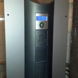 Szary piec Alpha Inno Tec z miedzianą instalacją i manometrem w pomieszczeniu z białymi płytkami. Widoczny panel sterowania z niebieskim wyświetlaczem.