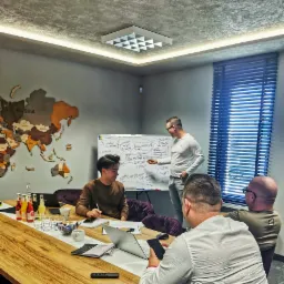 Prezentacja z zakresu PR dla Industry IMS: Mężczyzna w okularach tłumaczy zagadnienia na tablicy whiteboard grupie osób podczas spotkania w biurze. Na stole leżą laptopy, napoje i materiały.
