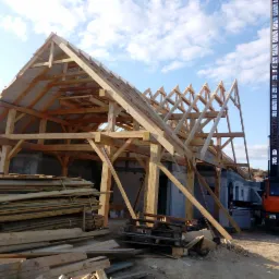 Wznoszenie drewnianej konstrukcji dachu na budynku z bloczków, widoczny dźwig samochodowy MAN i stos drewna budowlanego na placu budowy.