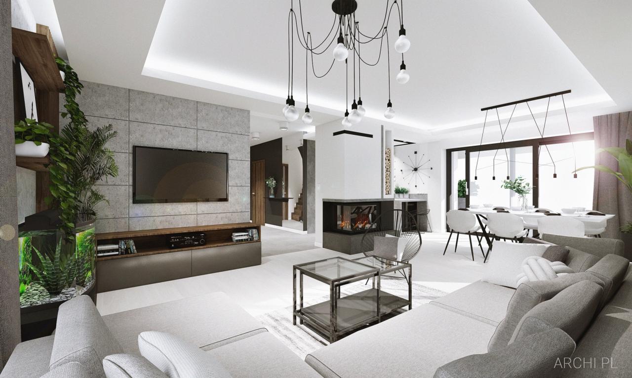Wnętrze przestronnego salonu z szarą sofą modułową, szklanym stolikiem kawowym, kominkiem i telewizorem na ścianie z betonowych płyt. Nad sofą zwisa nowoczesna lampa z żarówkami. W tle widoczna...