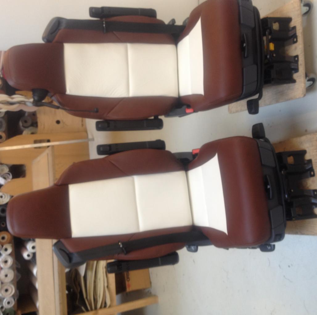 Dwa odnowione fotele samochodowe z brązowej skóry z białymi wstawkami, ustawione obok siebie na drewnianych paletach w warsztacie, widoczne rolki materiałów i półki w tle.