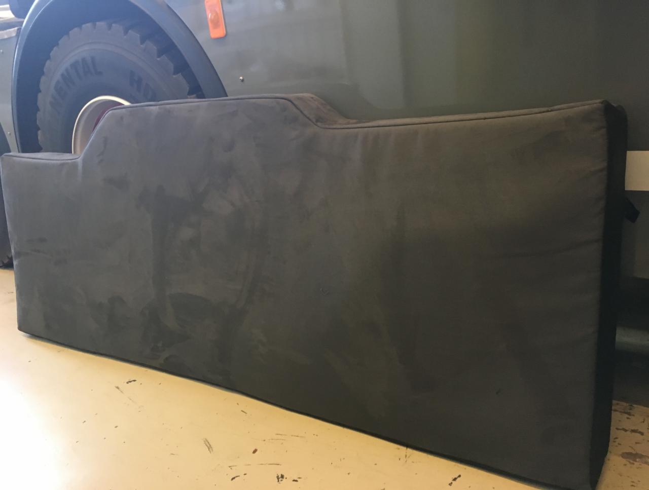 Szara, tapicerowana osłona, prawdopodobnie tylna ściana kabiny samochodu ciężarowego, leżąca na podłodze, z widocznym fragmentem opony pojazdu w tle.