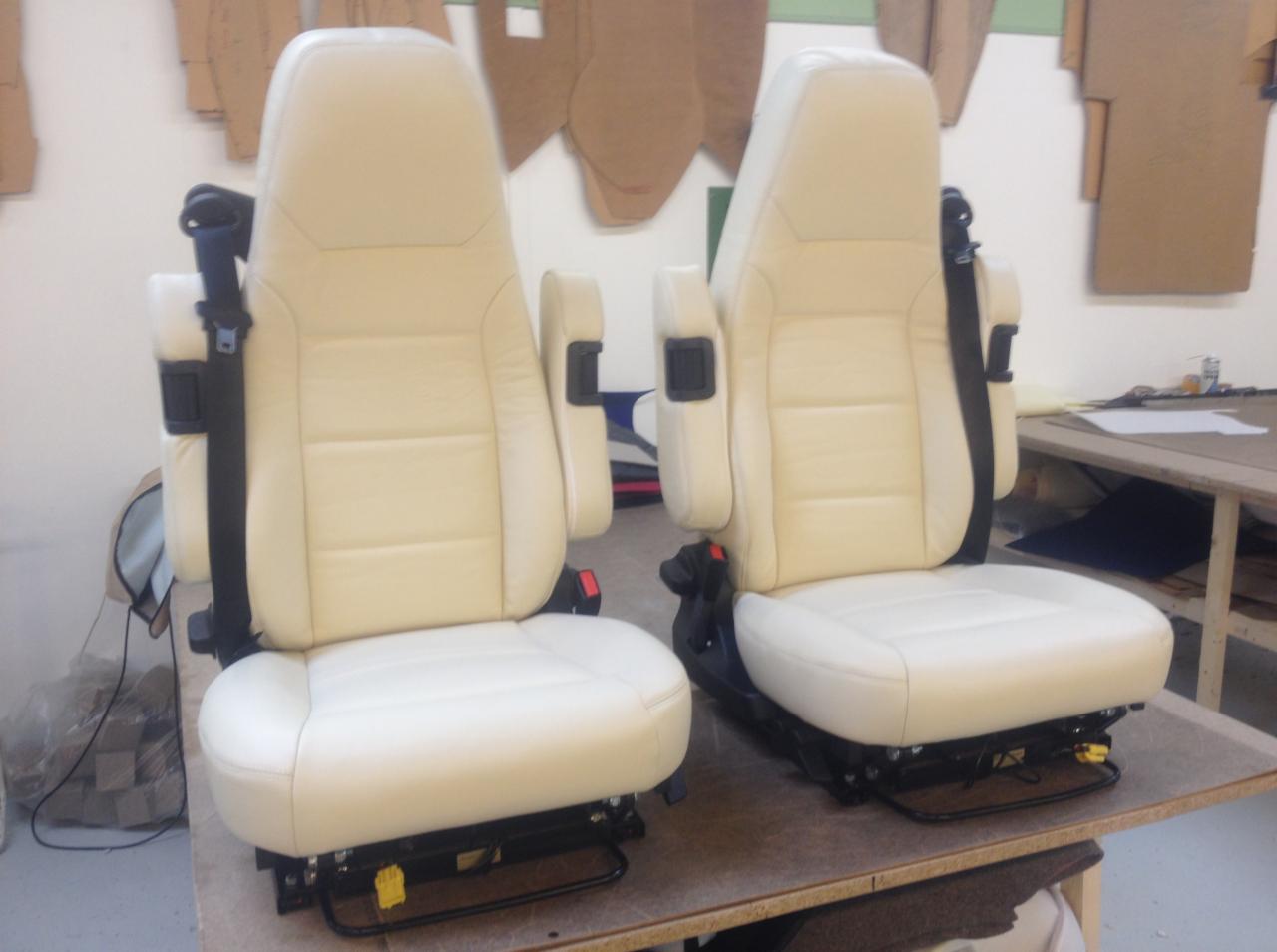 Dwa jasne, skórzane fotele samochodowe z pasami bezpieczeństwa i podłokietnikami, stojące na stole w warsztacie tapicerskim, z szablonami na ścianie w tle.
