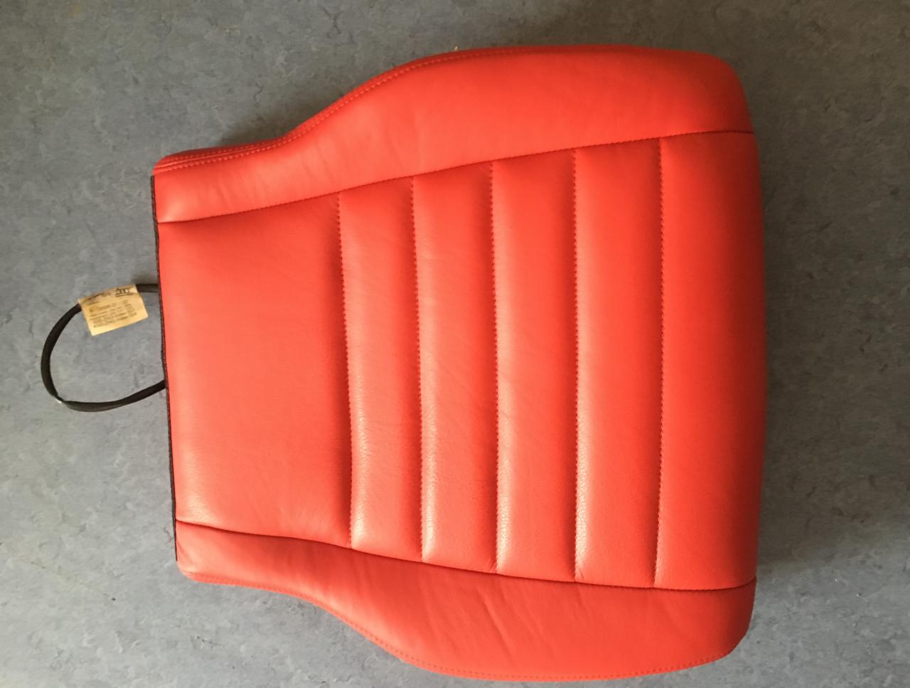 Czerwony, skórzany zagłówek samochodowy z pionowymi przeszyciami, leżący na szarym tle, z widocznym kablem i etykietą po lewej stronie.
