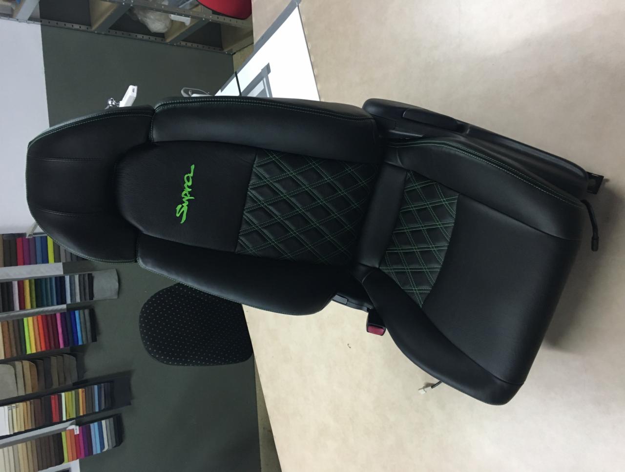 Odnowione czarne skórzane siedzenie samochodowe z zielonym logo 'Supra' i zielonymi szwami w romby, widoczne próbki materiałów tapicerskich w tle.