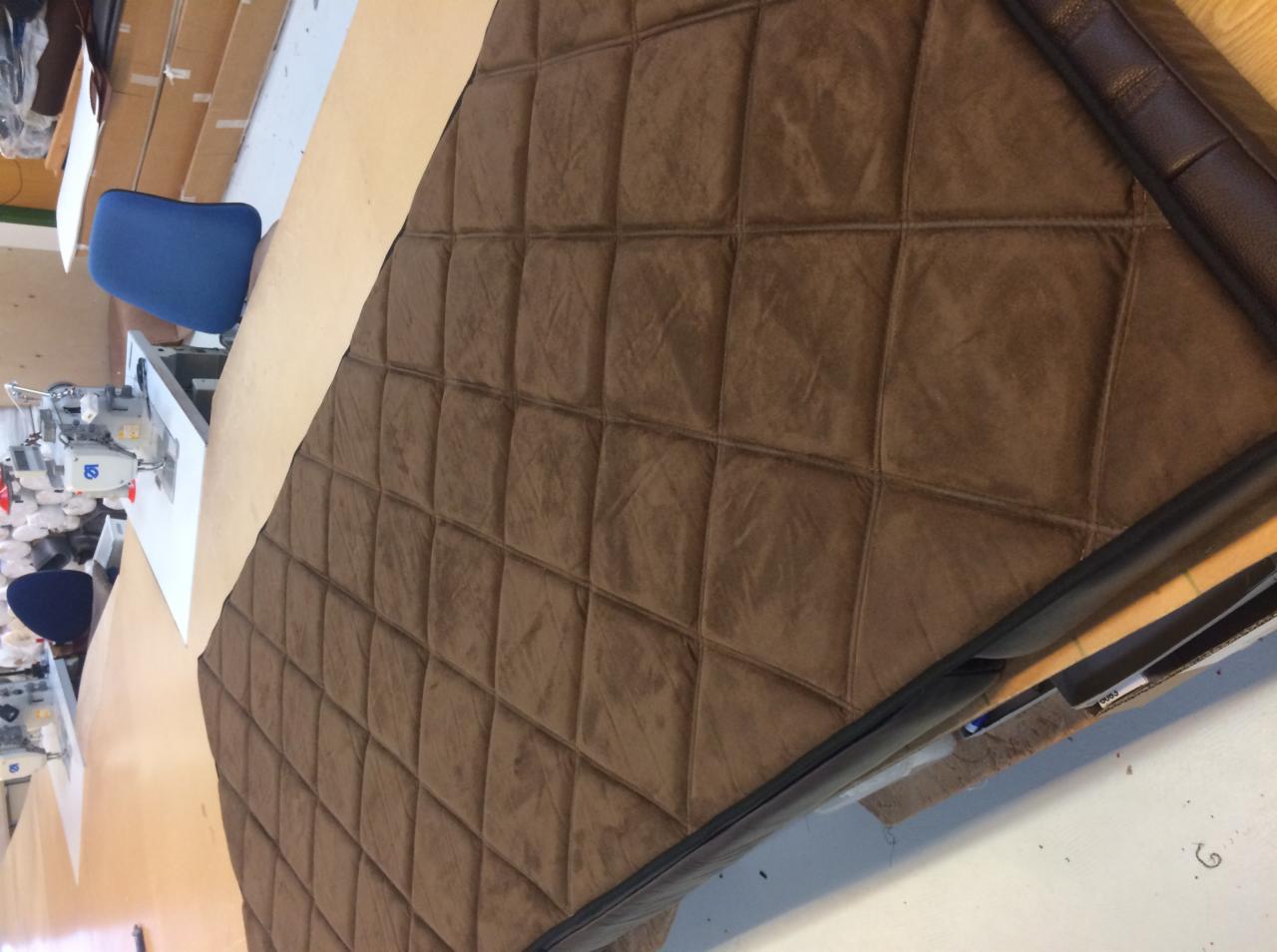 Brązowa, pikowana tapicerka samochodowa z ciemnobrązowym obramowaniem, leżąca na stole w warsztacie tapicerskim, obok maszyna do szycia i niebieskie krzesło.