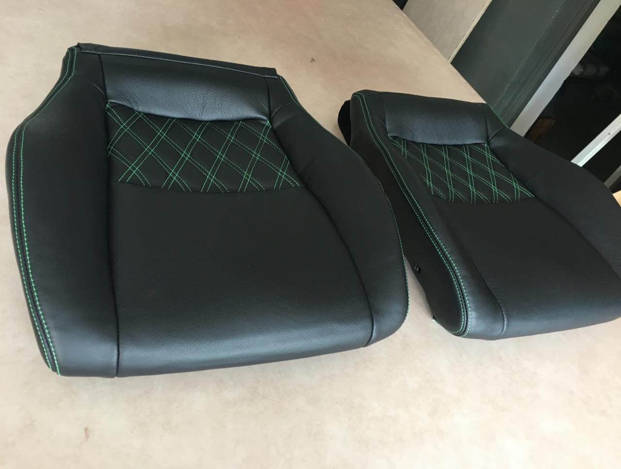 Dwa czarne, skórzane fotele samochodowe z zielonymi przeszyciami w romby, leżące na drewnianej powierzchni.