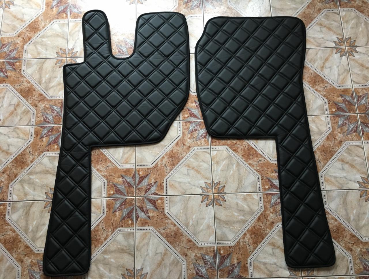 Dwa czarne, pikowane dywaniki samochodowe, leżące na kafelkach w odcieniach beżu i brązu.