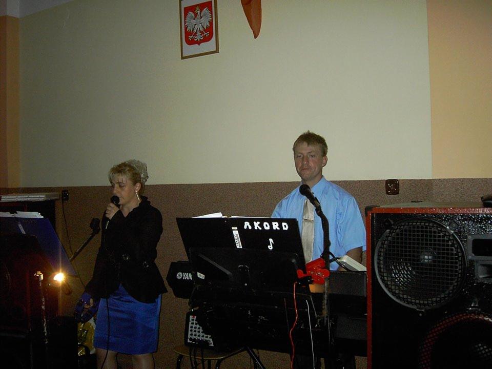 Duet muzyczny: kobieta z mikrofonem i mężczyzna grający na keyboardzie z napisem 'Akord'. W tle godło Polski.