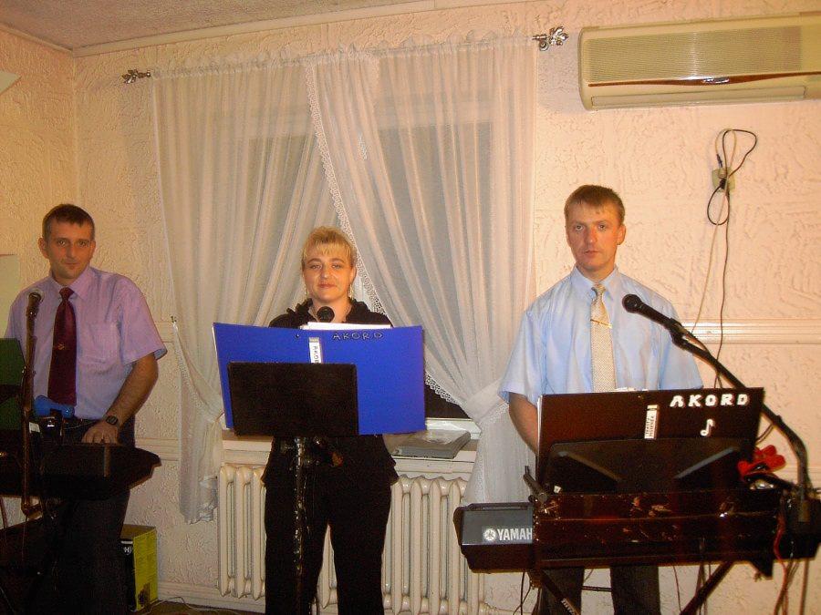 Zespół muzyczny Akord w trakcie występu: troje muzyków gra na keyboardach Yamaha, za nimi białe zasłony i klimatyzator. Jeden z muzyków ma mikrofon.