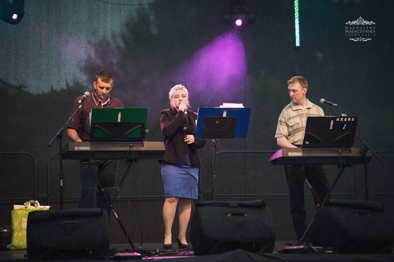 Zespół muzyczny 'Akord' w trakcie występu na scenie. Wokalistka śpiewa do mikrofonu, a dwaj klawiszowcy grają na instrumentach Yamaha. Scena oświetlona fioletowym światłem.