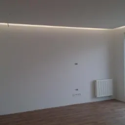 Wnętrze pokoju po remoncie z białymi ścianami, drewnopodobną podłogą i oknem balkonowym, widoczne ukryte oświetlenie LED w suficie podwieszanym.