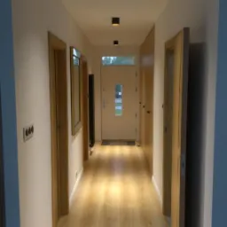 Korytarz po remoncie z drewnopodobną podłogą, białymi ścianami i trzema czarnymi lampami sufitowymi.