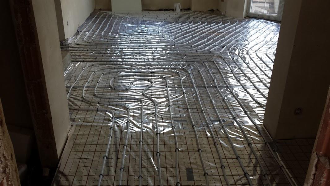 Instalacja ogrzewania podłogowego z rur na folii, widok z góry w surowym wnętrzu. Szare rury ułożone spiralnie na siatce, przygotowane do zalania wylewką.