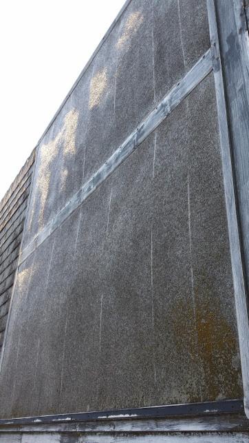Szara ściana budynku z widocznymi śladami zacieków i zabrudzeń, fragment dachu i metalowe elementy konstrukcyjne. Widok z dołu, perspektywa zniekształcona.