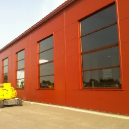 Czerwona hala z panelami warstwowymi i oknami. Żółty podnośnik koszowy stoi obok budynku. Widoczne łączenia paneli i faktura elewacji.